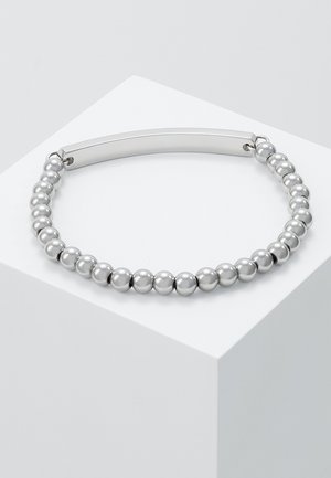 Bracelet en argent avec perles et une plaque rectangulaire lisse sur une surface géométrique blanche.