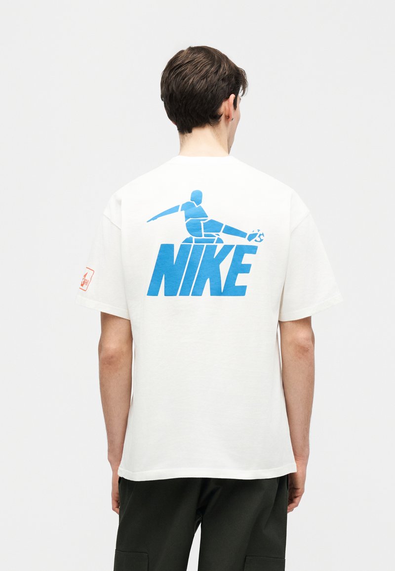 Junger Mann trägt ein weißes T-Shirt mit einem blauen Bild eines Fußballspielers, der über dem Wort "NIKE" auf der Rückseite einen Ball tritt.