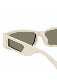 Occhiali da sole color crema con montatura angolare a forma di occhio di gatto, lenti grigie e steli affusolati con un piccolo dettaglio rettangolare accentato.