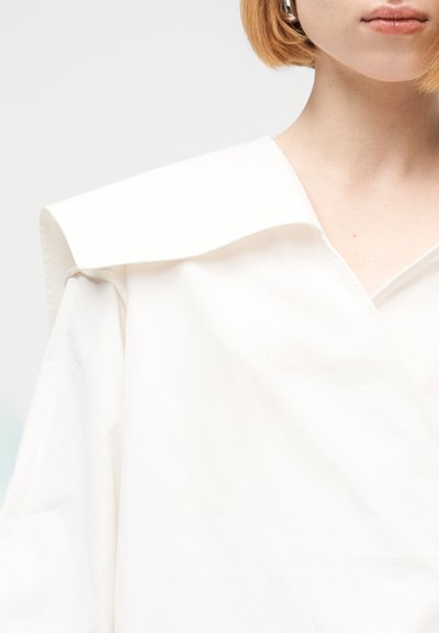 Κοντινό πλάνο ενός ατόμου με κοντά κόκκινα μαλλιά που φοράει λευκό ρούχο με φαρδιά γιακά και V-λαιμόκοψη.