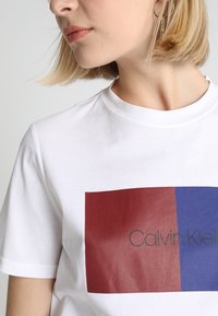 Bílý bavlněný tričko s obdélníkovou grafikou v tmavě červené a modré barvě, se středově umístěným logem "Calvin Klein" v černé barvě. Krátké rukávy.