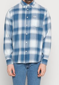 Chemise à carreaux bleue et blanche en tissu léger, avec une poche poitrine et un col classique.