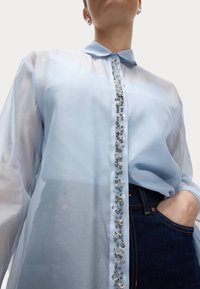 Blusa trasparente azzurro chiaro con colletto, caratterizzata da una placca con bottoni decorativi impreziosita da gemme e perline, abbinata a jeans scuri.
