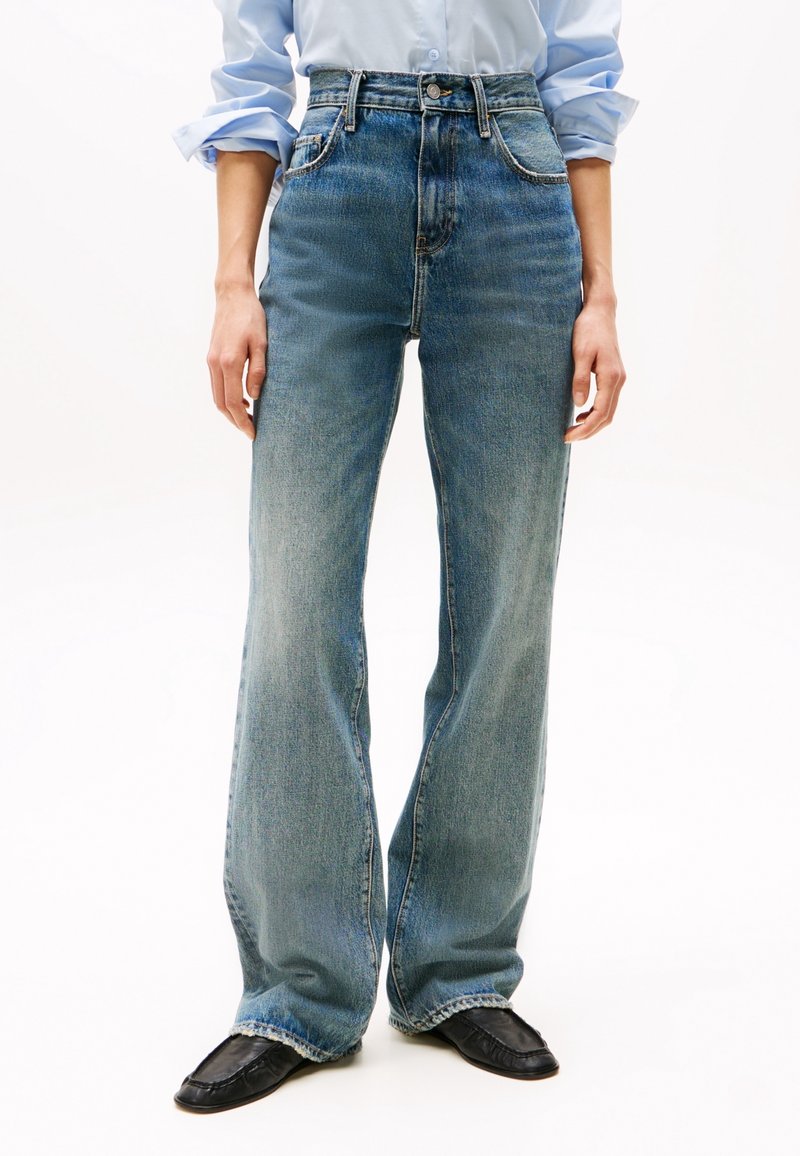 Personne portant un jean taille haute, délavé clair et à jambes larges, associé à des chaussures noires à bout carré et une chemise bleu clair boutonnée avec manches retroussées.