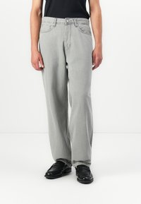 Person som har på seg løse lysegrå jeans sammen med svarte loafers og en svart topp, stående mot en ensfarget hvit bakgrunn.
