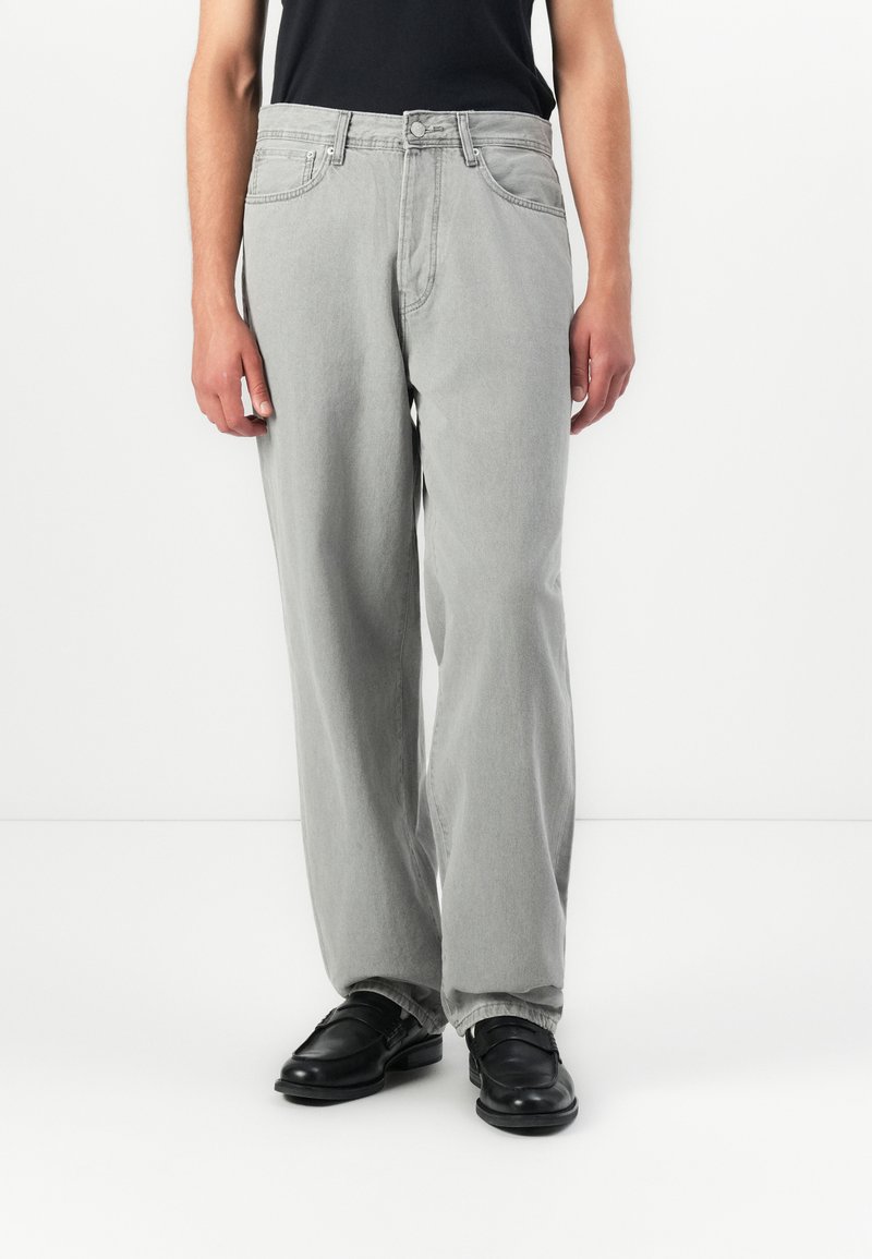 Person som har på seg løse lysegrå jeans sammen med svarte loafers og en svart topp, stående mot en ensfarget hvit bakgrunn.