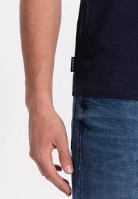 Námořnické modré bavlněné tričko s krátkým rukávem, zobrazené z boku, s etiketou na lemu. Nosené s texturovanými modrými džínami.