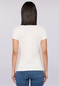 Bílý tričko s krátkým rukávem, kulatým výstřihem, přiléhavým střihem a hladkou strukturou. Nosené s tmavými džíny.