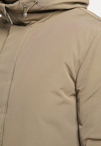 Béžová zateplená bunda s matným povrchem, vybavená vysokým límcem, kapucí se stahovací šňůrkou a viditelnými švy podél švů pro odolnost.