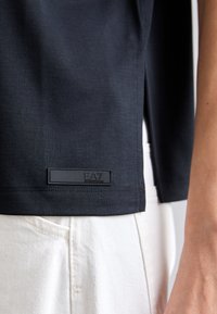 Mustat ribbed lyhythihaiset paita, jossa on sivurei'ät, tekstuuri pinta ja suorakulmainen EA7-logo helmaassa. Yhdistetty valkoisten housujen kanssa.
