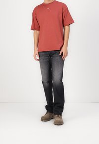 Röd t-shirt med kort ärm, grå jeans och beige mockaskor. T-shirten har en liten logotyp på bröstet; jeansen har en avslappnad passform.