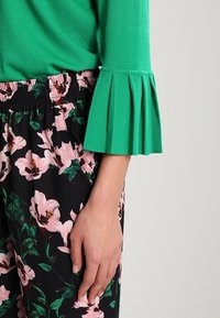 Haut en tricot vert avec manches trois-quarts plissées, associé à un pantalon noir avec un motif floral rose et une ceinture élastique.