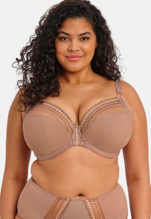 Frau mit lockigem Haar trägt einen hautfarbenen BH und passende hoch taillierte Unterwäsche mit gemustertem Netzstoff und lächelt in die Kamera.