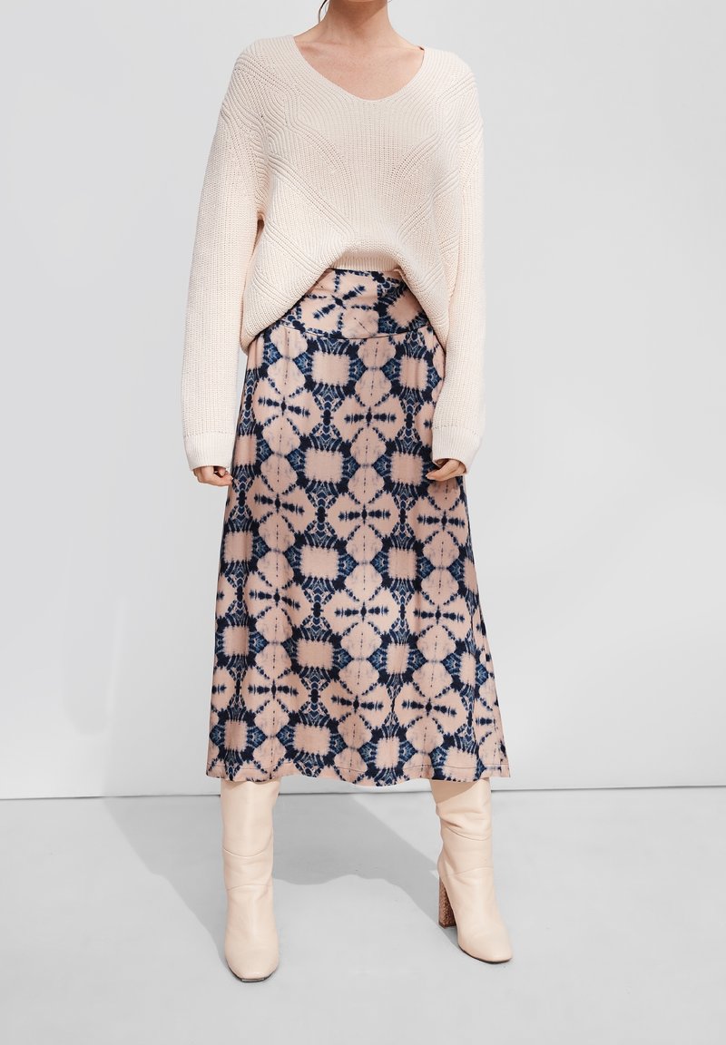 Lichtroze gebreide trui met een V-hals, gecombineerd met een lange gemUSTERDE rok in roze en blauwe tinten, met geometrische ontwerpen. Beige enkellaarsjes.