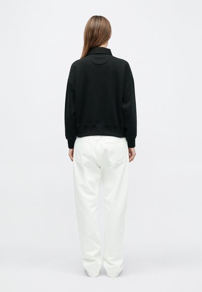 Svart genser med høy krage og ribbestrikkede mansjetter, kombinert med løstsittende hvite bukser med baklommer.