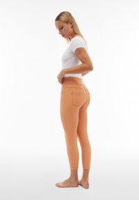 Leggings color pesca con un materiale liscio e elastico, dotati di una vita alta e tasche posteriori, visti di lato.