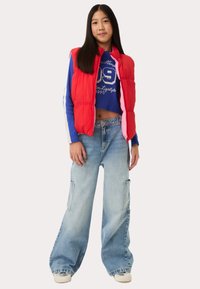 Rode puffer vest over een blauwe cropped sweatshirt, losse lichtblauwe jeans met zijzakken, en witte sneakers. Strakke lijnen en casual fit.