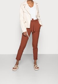 Giacca in ecopelle beige chiaro, t-shirt bianca, pantaloni tapered color ruggine con cintura e sandali chiari con dettagli intrecciati.