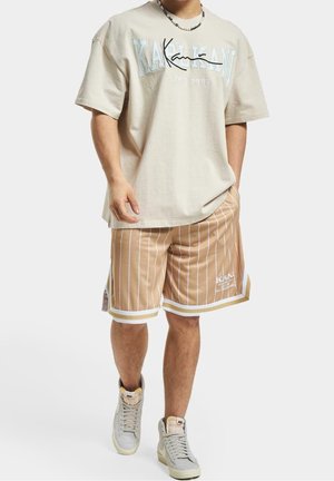 T-shirt beige oversize avec lettrage bleu et noir, associé à un short beige à rayures. Accents blancs à l'ourlet, porté avec des baskets montantes gris clair.