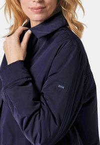 Marineblaue, leichte Jacke mit weitem Kragen, glatter Textur und subtiler Markenpatch am Ärmel.