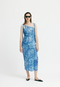 Blaues, gerafftes Kleid mit einseitigem Design, aus strukturiertem Stoff und leichten Seitenschlitzen. Getragen mit schwarzen flachen Schuhen und Sonnenbrille.