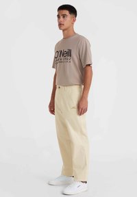 T-shirt beige avec logo noir imprimé, associé à un pantalon jaune clair et des baskets blanches avec des accents noirs. Lignes épurées, coupe décontractée.