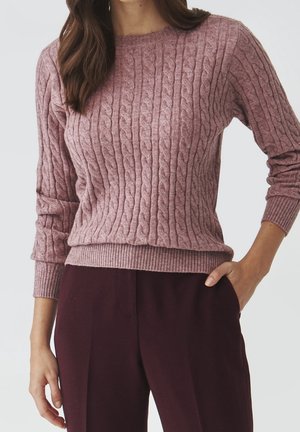Femme portant un pull en maille torsadée mauve avec manches longues et poignets côtelés, assorti à un pantalon bordeaux foncé, une main dans la poche.