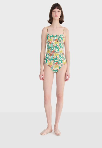 Femme portant un maillot de bain une pièce à fines bretelles et motif floral coloré, debout devant un fond clair uni.
