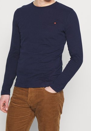 Marineblaue Langarmshirt aus weicher Baumwolle mit Rundhalsausschnitt und kleinem Logo auf der Brust. Kombiniert mit braunen Cordhosen.