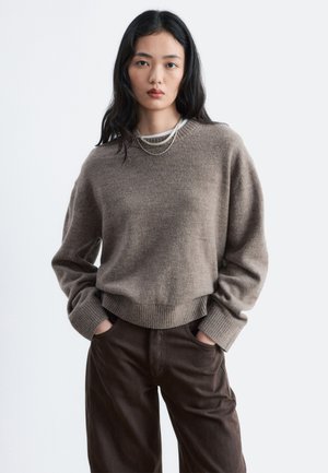 Jeune femme aux longs cheveux noirs portant un pull couleur taupe, des colliers superposés et un pantalon marron foncé, les mains dans les poches.