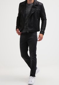 Schwarze Leder-Bikerjacke mit Reißverschluss, strukturiertes graues Strickpullover, dunkle Hose und wei�ße Sneakers. Das Modell steht vor einem neutralen Hintergrund.
