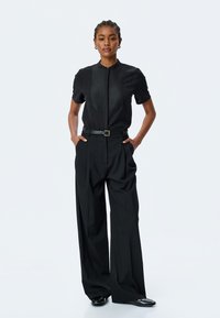 Blusa negra de manga corta con un panel texturizado y cremallera en el frente, combinada con pantalones negros de talle alto y pliegues, y un cinturón negro ajustado.
