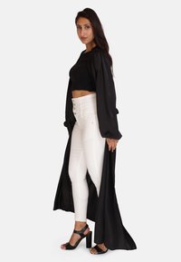 Kvinde stående i hvide, højtaljede, knapede bukser, sort crop top, lang sort åben frakke og sorte højhælede sandaler, mod hvid baggrund.