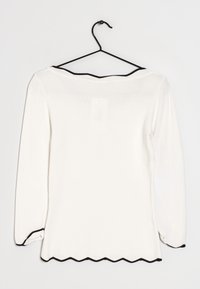 Camicia bianca a maniche lunghe esposta su una gruccia nera, con un bordo nero scallopato lungo il collo e i polsini.