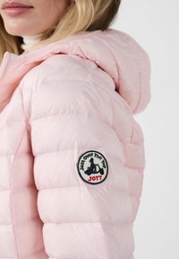 Lichtroze donsjack met verticale doorgestikte naden en een capuchon met trekkoord, voorzien van een ronde patch met zwarte, witte en rode tekst op de mouw.