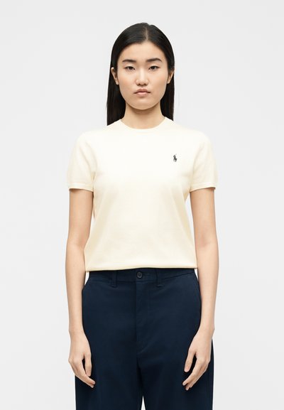 Νεαρή γυναίκα με ίσια μαύρα μαλλιά, που φοράει κρεμ κοντομάνικο πουκάμισο με ένα μικρό λογότυπο και σκούρο μπλε παντελόνι, στέκεται μπροστά σε απλό φόντο.