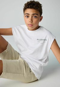 Biely bezrukávový tričko s logom "ALLSAINTS" v zelenej farbe, v kombinácii s béžovými šortkami so subtilným textom. Hladká textúra látky.