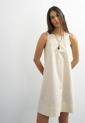 Femme aux longs cheveux foncés portant une robe beige sans manches et un long collier avec un pendentif foncé, debout devant un fond clair uni.