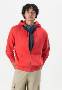 Crvena zip-up dukserica s kengurua džepom i kapuljačom na šnurlanje. Nosena preko tamnoplave majice s plavom maramom oko vrata. Ogrtači od perli.