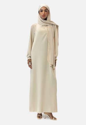 Long, beige gown made of smooth fabric, featuring long sleeves, cuff details, and a matching scarf draped elegantly. High-heeled sandals complete the look.