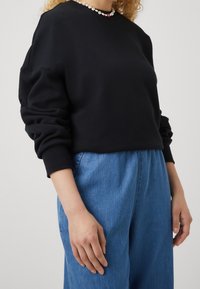 Persona che indossa una felpa nera a maniche lunghe, jeans a vita alta blu e una collana di perline colorate su uno sfondo neutro.
