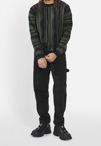 Strickpullover in Dunkelgrün und Schwarz mit vertikalen Mustern, kombiniert mit schwarzen Hosen und klobigen schwarzen Sneakers.