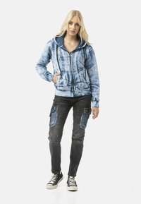 Blue tie-dye zip-up hoodie with a contrasting dark blue hood, paired with black cargo pants featuring denim accents and pockets. Black sneakers.