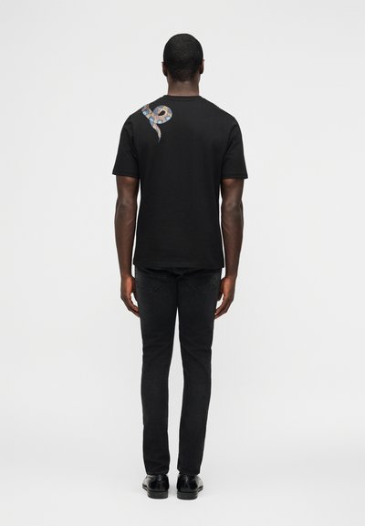 Άνδρας που στέκεται με την πλάτη προς τον θεατή, φορώντας μαύρο μπλουζάκι με μικρό μπλε και μπεζ σχέδιο φιδιού στον δεξί ώμο, μαύρο τζιν και μαύρα παπούτσια.