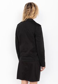 Giacca di jeans nera con spalline e polsini con zip. Presenta una texture opaca liscia e una vestibilità sartoriale.