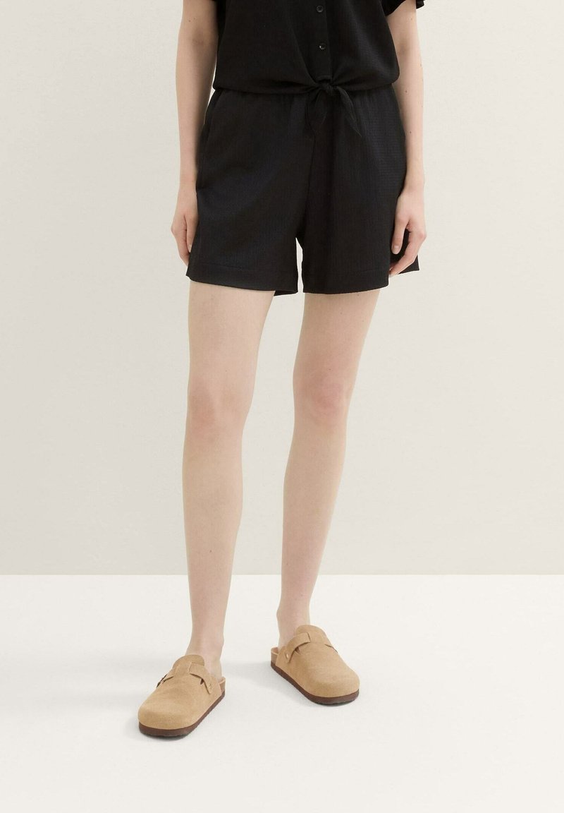 Personne portant un short noir ample et des chaussures à enfiler couleur sable, debout devant un fond uni clair.