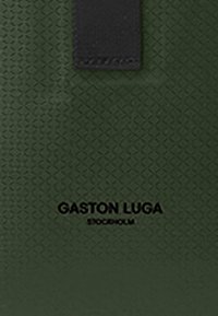 Grønt strukturert materiale med en diamantmønster. Produktet har et svart lærakcent med "GASTON LUGA STOCKHOLM" trykket i svart.