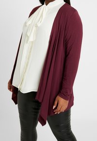 Femme portant une blouse blanche à volants, un cardigan ouvert bordeaux foncé et un pantalon en cuir noir, se tenant devant un fond clair.