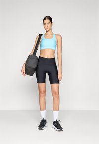 Sujetador deportivo azul claro con logo, pantalones cortos de compresión negros de cintura alta, zapatillas deportivas negras y una bolsa de hombro gris, sobre un fondo liso.