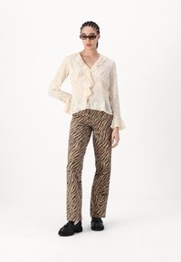 Helle, halbtransparente Cremebluse mit Rüschendetails, kombiniert mit braunen, tigerstreifenmuster hohen Hosen und schwarzen klobigen Schuhen.